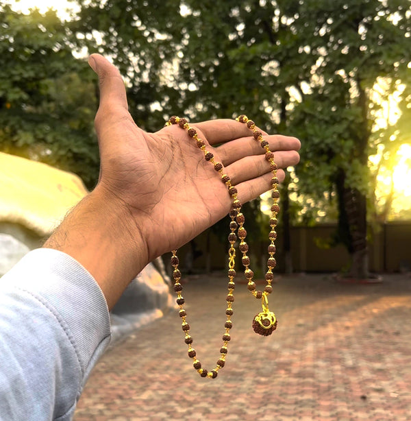 Gold-plated rudraksha Om Shree mala held in hand outdoors at golden hour, showing full necklace length
