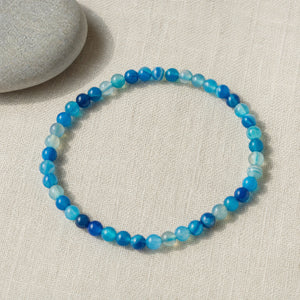 Soultheory Blue Agate 4mm Round Bead Bracelet on beige linen beside a river pebble