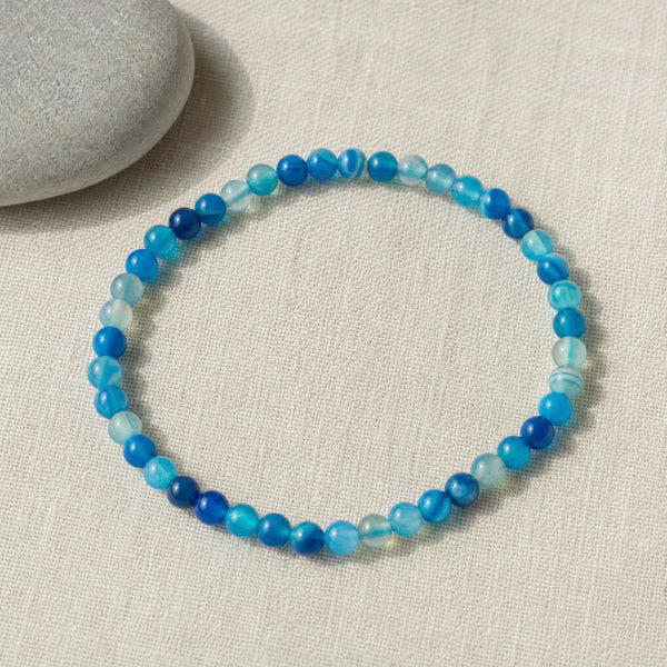 Soultheory Blue Agate 4mm Round Bead Bracelet on beige linen beside a river pebble