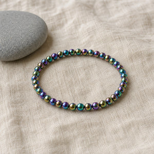 Soultheory Rainbow Hematite 4mm Bead Bracelet on beige linen beside a river pebble