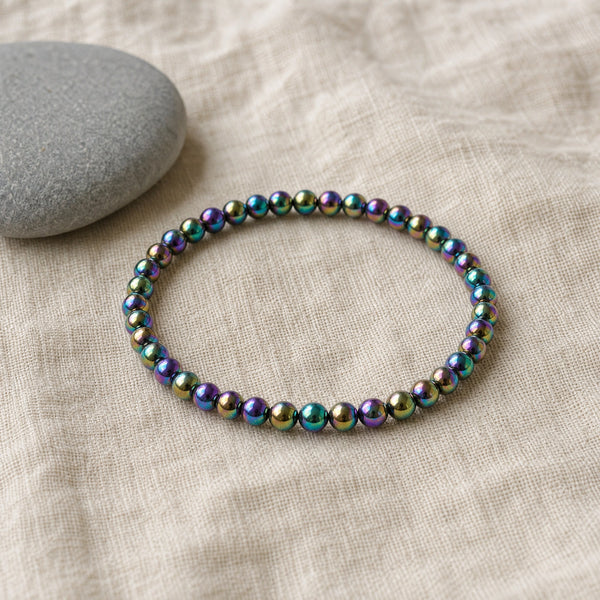 Soultheory Rainbow Hematite 4mm Bead Bracelet on beige linen beside a river pebble