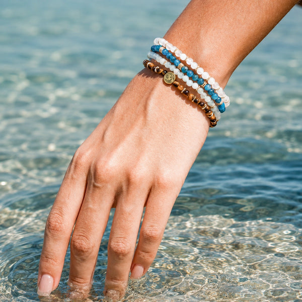 Four dainty 4 mm Abundance gemstone bracelets with Soultheory's gold charm worn beside sunlit turquoise water
