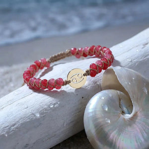 Cherry pink Cancer zodiac bracelet resting on beach driftwood beside a seashell, gold crab sign charm