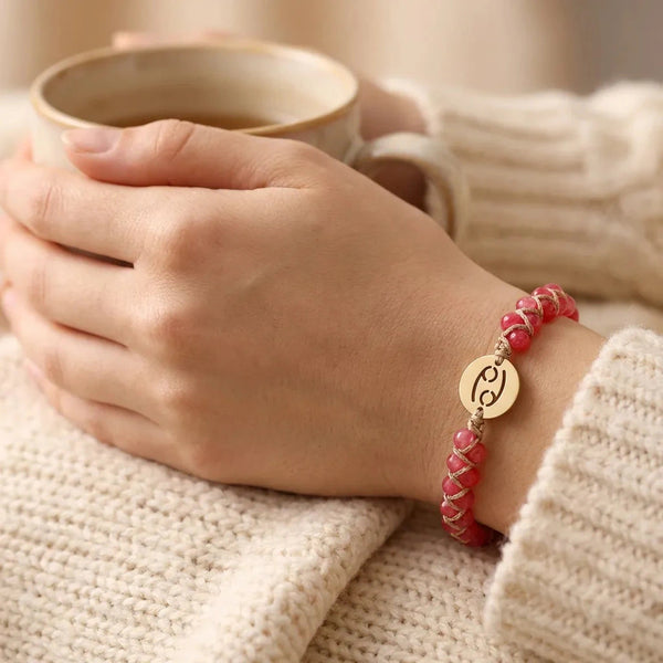 Cherry pink Cancer zodiac bracelet worn on a woman's wrist holding a warm mug, gold crab sign charm on top