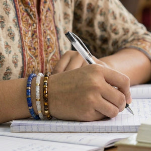 Student wearing clear quartz, tiger eye and lapis lazuli bracelet stack while writing exam notes for focus